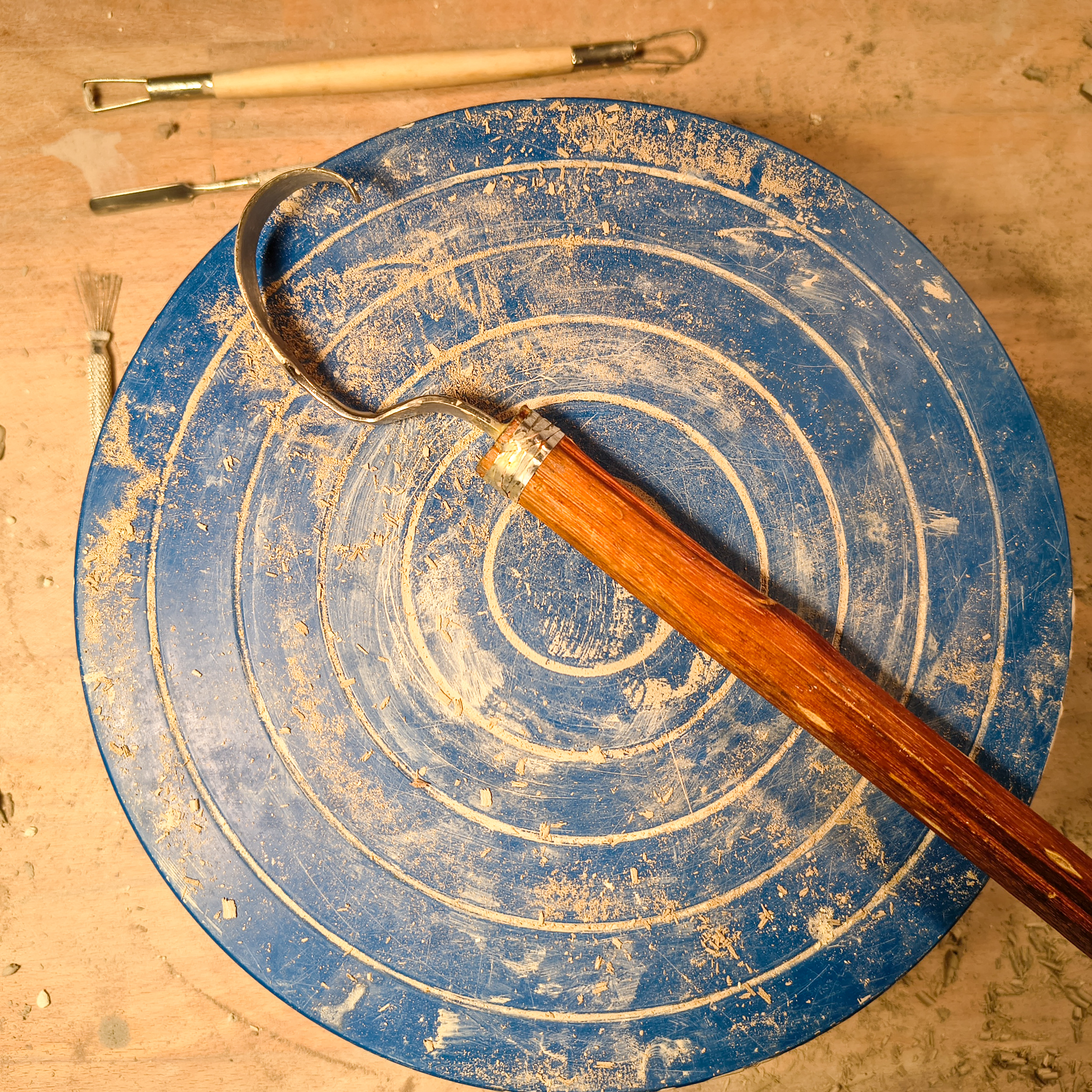 A blue pottery wheel with clay residue and spiral marks from use. Pottery tools lie next to it on a wooden surface.