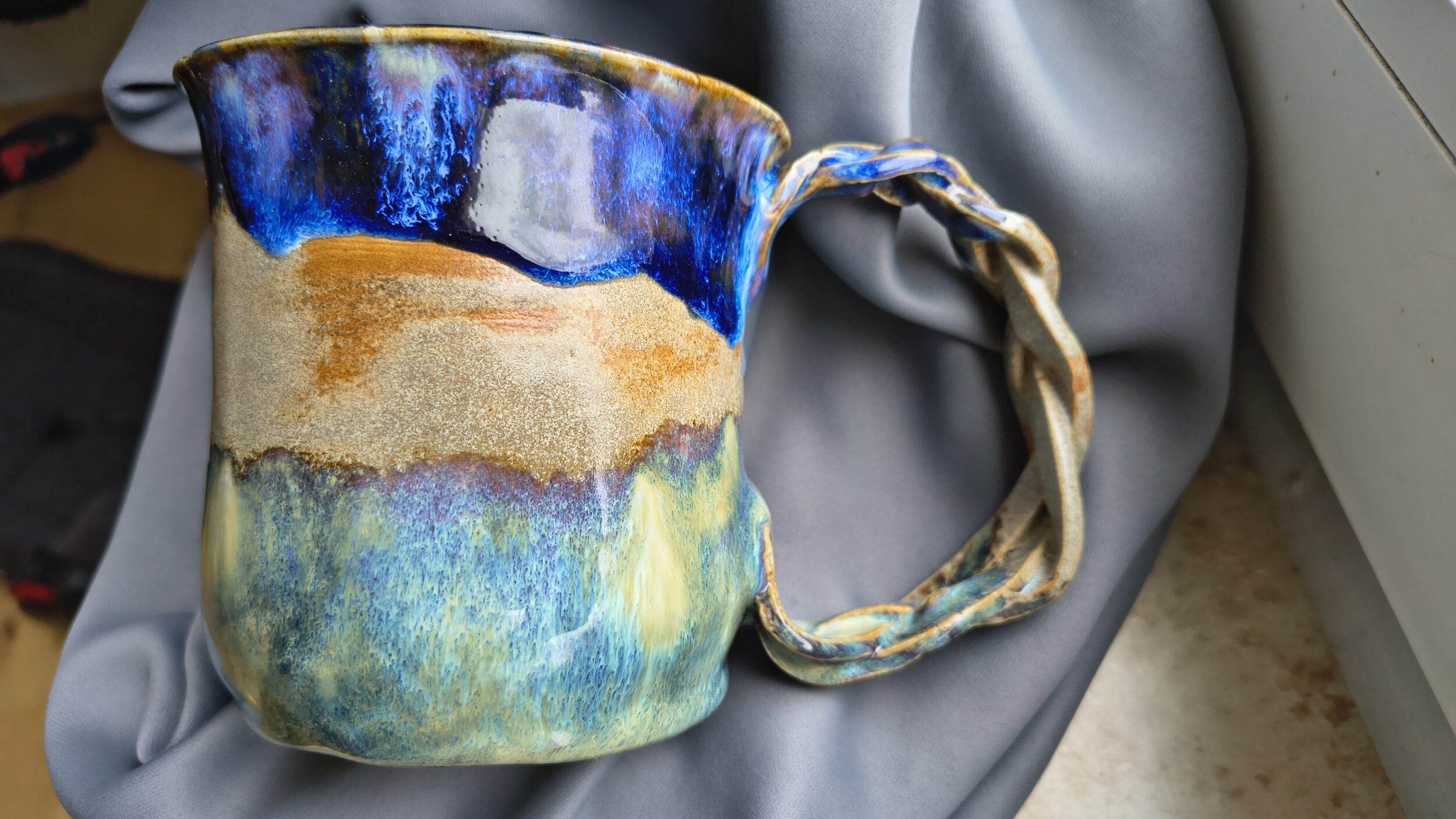 A handmade ceramic cup with multi-colored glaze in shades of blue, beige, and green lies on gray fabric. The cup has